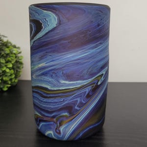 May include: A cylindrical vase with a swirling pattern of blues, purples, and hints of gold. The design resembles a watercolor painting or the flow of water. The vase is on a dark surface, with a green plant in the background.