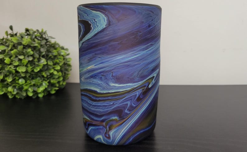 May include: A cylindrical vase with a swirling pattern of blues, purples, and hints of gold. The design resembles a watercolor painting or the flow of water. The vase is on a dark surface, with a green plant in the background.