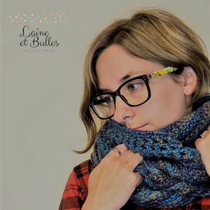 May include: A woman wearing a red and black checked shirt and a blue and brown crocheted infinity scarf. The scarf is folded over and the woman is holding it with her right hand. The woman is looking to the left of the image. The text "Laine et Bulles" is visible in the top left corner of the image.