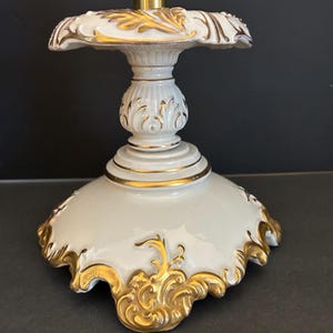 May include: A cream-colored, ornate pedestal with gold accents. The base and top feature intricate gold scrollwork and trim. The pedestal has a fluted column and a scalloped edge, creating an elegant, vintage aesthetic.