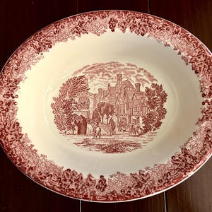 May include: Oval serving bowl with a cream-colored interior and a burgundy border featuring a grape and leaf design. The center showcases a detailed burgundy illustration of a historic building with figures.