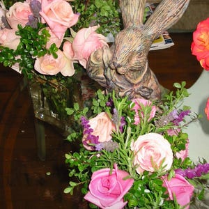 May include: A brown rabbit figurine with long ears is surrounded by a bouquet of pink roses and lavender flowers in a clear glass vase. The flowers are arranged in a rustic wooden planter.