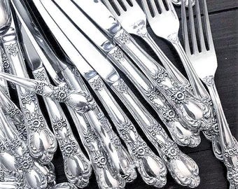 May include: A collection of silver-toned flatware featuring ornate floral and scrollwork detailing on the handles. The set includes multiple knives and forks arranged in a cluster, showcasing polished metal surfaces with intricate embossed patterns.