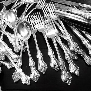 May include: A collection of polished silver-toned flatware featuring ornate, embossed floral detailing on the handles. The set includes multiple forks with four tines, rounded spoons, and knives with smooth blades, all arranged in a dense, overlapping cluster.