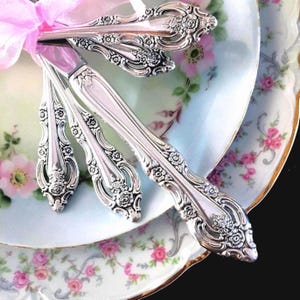 May include: A set of three silver-toned metal spoons featuring an ornate floral embossed pattern on the handles, tied together with a light pink ribbon. The spoons are arranged in a fan shape, showcasing detailed scrollwork and rose motifs.