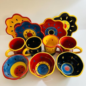 May include: A collection of ceramic tableware featuring four scalloped plates and seven mugs and bowls. The pieces are decorated with vibrant patterns in red, yellow, blue, and black, including circular motifs, dots, and dashes on each item.
