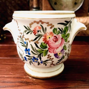 May include: A white ceramic planter with a floral design. The planter features a base and two handles. The floral design includes pink roses, blue and purple flowers, and green leaves, with gold trim.
