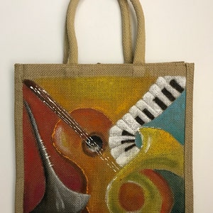 May include: A jute tote bag featuring a painted design of musical instruments. The design includes a guitar, a trumpet, and a keyboard in shades of yellow, orange, red, blue, and white.