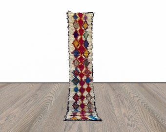 May include: A long, narrow, white rug with a colorful diamond pattern in shades of red, blue, yellow, and purple. The rug has a fringe border.