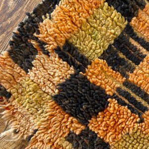 May include: Close-up of a colorful, hand-woven rug with a geometric pattern in shades of orange, black, and brown. The rug has a thick, textured pile and a frayed edge.