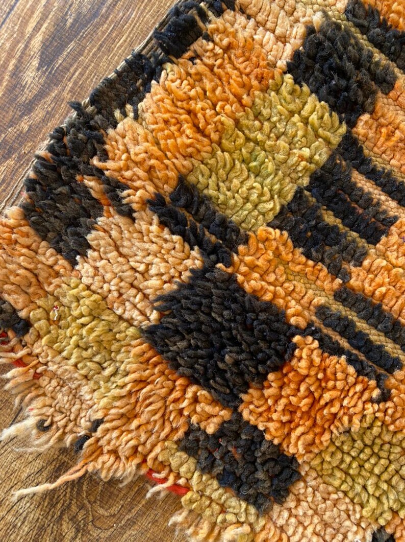 May include: Close-up of a colorful, hand-woven rug with a geometric pattern in shades of orange, black, and brown. The rug has a thick, textured pile and a frayed edge.