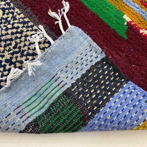 May include: Close-up of a colorful woven rug with blue, green, red, yellow, and white stripes. The rug has a fringe border and is made of a soft, textured material.