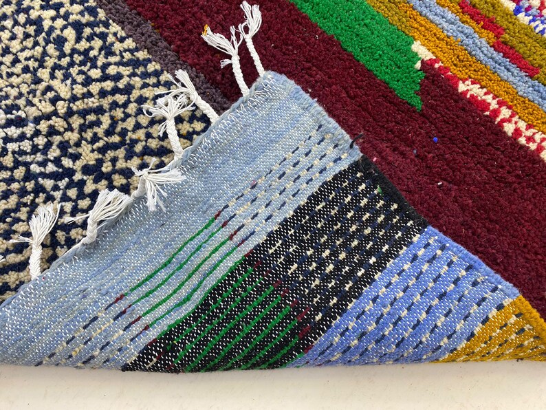 May include: Close-up of a colorful woven rug with blue, green, red, yellow, and white stripes. The rug has a fringe border and is made of a soft, textured material.
