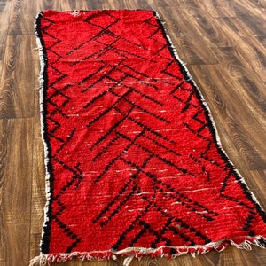 Op de afbeelding: Een rood en zwart Marokkaans tapijt met een geometrisch patroon. Het tapijt heeft een franje rand en is gemaakt van wol.