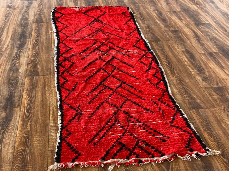 Op de afbeelding: Een rood en zwart Marokkaans tapijt met een geometrisch patroon. Het tapijt heeft een franje rand en is gemaakt van wol.