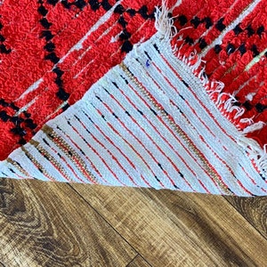 Op de afbeelding: Close-up van de achterkant van een rood en zwart tapijt met witte franjes. Het tapijt is omgeslagen om de witte achterkant met rode en zwarte strepen te laten zien.