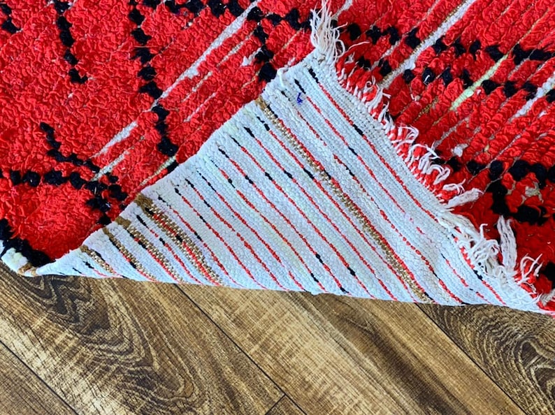 Op de afbeelding: Close-up van de achterkant van een rood en zwart tapijt met witte franjes. Het tapijt is omgeslagen om de witte achterkant met rode en zwarte strepen te laten zien.