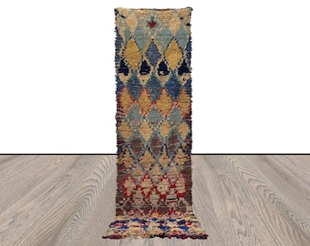 May include: A long, narrow, handwoven rug with a geometric pattern in shades of blue, red, yellow, and brown. The rug has a fringed edge and is made of wool.