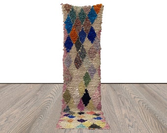 May include: A long, narrow rug with a pink background and a diamond pattern in shades of blue, green, orange, and purple. The rug is made of wool and has a hand-woven texture.