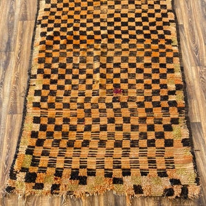 May include: A vintage Moroccan rug with a checkered pattern in shades of orange and black. The rug has a worn and distressed look, adding to its unique character.