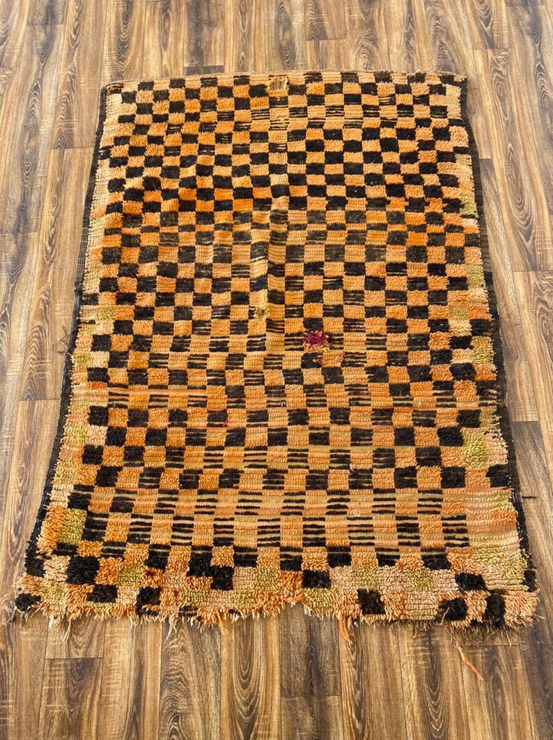May include: A vintage Moroccan rug with a checkered pattern in shades of orange and black. The rug has a worn and distressed look, adding to its unique character.