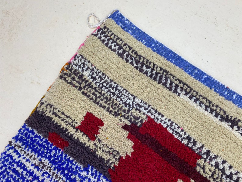 May include: A colorful striped rug with a geometric pattern in shades of red, blue, white, and gray. The rug is made of a soft, fluffy material and has a fringe border.