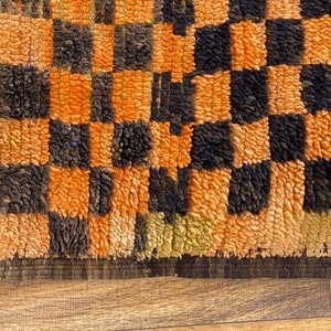 May include: Close-up of a woven rug with a checkered pattern in orange and black. The rug has a thick, fuzzy texture.