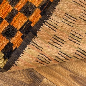 May include: Close-up of a woven rug with a black, orange, and beige geometric pattern. The rug is folded in half, revealing the underside of the rug.