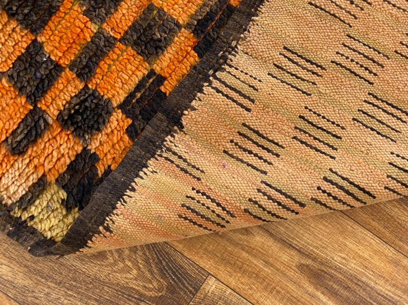 May include: Close-up of a woven rug with a black, orange, and beige geometric pattern. The rug is folded in half, revealing the underside of the rug.
