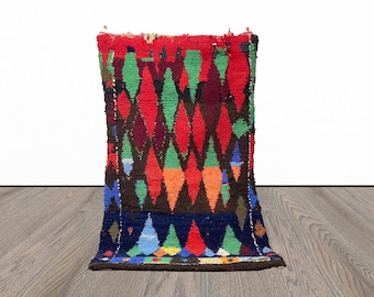 May include: A colorful Moroccan rug with a diamond pattern in shades of red, green, blue, and black. The rug is woven with a thick, textured yarn and has a fringe on the ends.