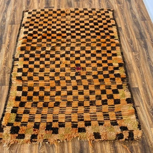 May include: A vintage Moroccan rug with a checkered pattern in shades of orange and brown. The rug has a frayed edge and is made of wool.