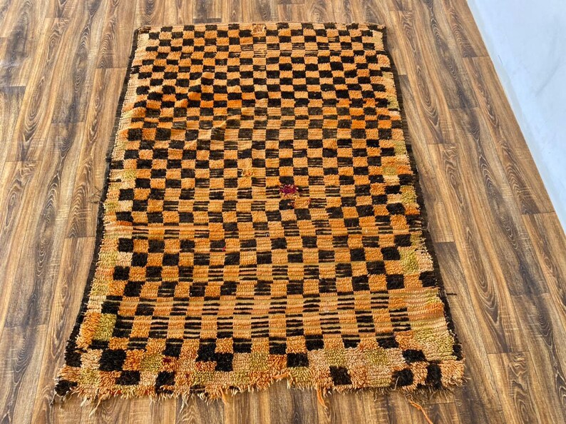 May include: A vintage Moroccan rug with a checkered pattern in shades of orange and brown. The rug has a frayed edge and is made of wool.