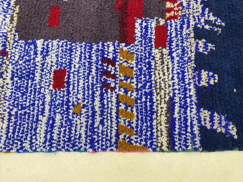 May include: A close-up of a colorful Moroccan rug with blue, white, red, brown, and gold geometric patterns. The rug has a thick, textured pile.