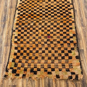 May include: A vintage Moroccan rug with a checkered pattern in shades of orange and black. The rug has a frayed edge and a worn, distressed look.