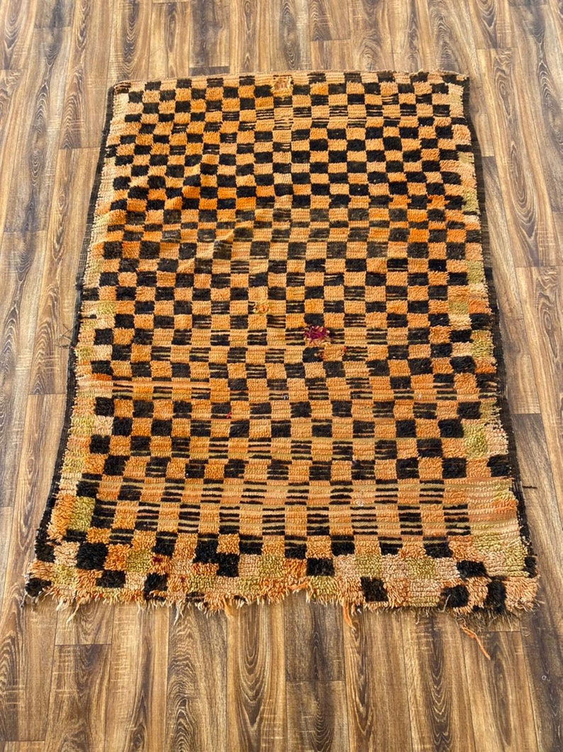 May include: A vintage Moroccan rug with a checkered pattern in shades of orange and black. The rug has a frayed edge and a worn, distressed look.