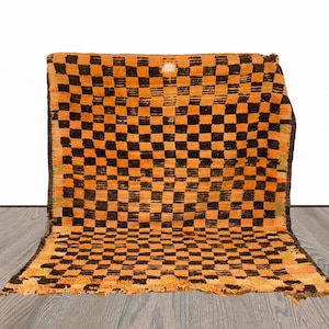 May include: A vintage orange and black checkered rug with frayed edges. The rug is folded in half and placed on a wooden floor.