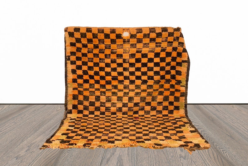 May include: A vintage orange and black checkered rug with frayed edges. The rug is folded in half and placed on a wooden floor.
