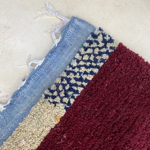 May include: A close-up of a colorful woven rug with a blue, white, and red pattern. The rug has a fringe border and is made of soft, textured fibers.