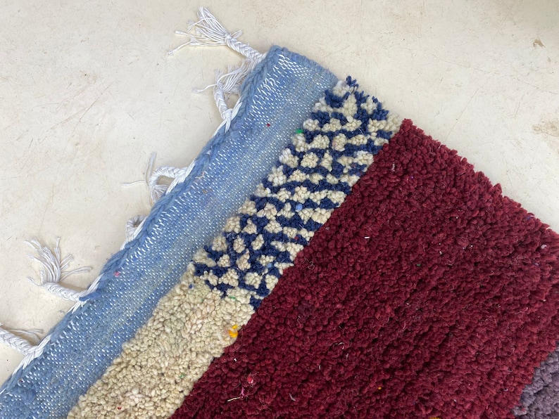 May include: A close-up of a colorful woven rug with a blue, white, and red pattern. The rug has a fringe border and is made of soft, textured fibers.