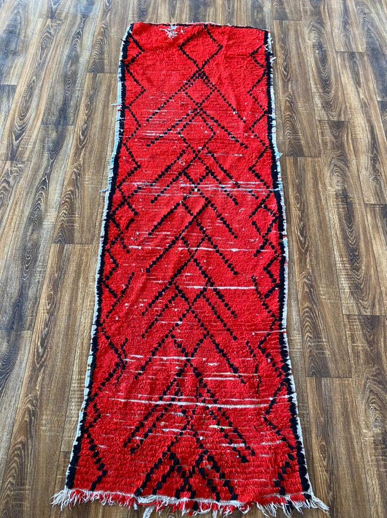 Op de afbeelding: Een rood en zwart Marokkaans tapijt met een ruitpatroon. Het tapijt heeft een franje rand en is gemaakt van wol.