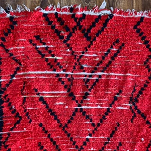 Op de afbeelding: Rood en zwart gestreept tapijt met een gefranst randje. Het tapijt heeft een geometrisch ontwerp met een witte achtergrond.
