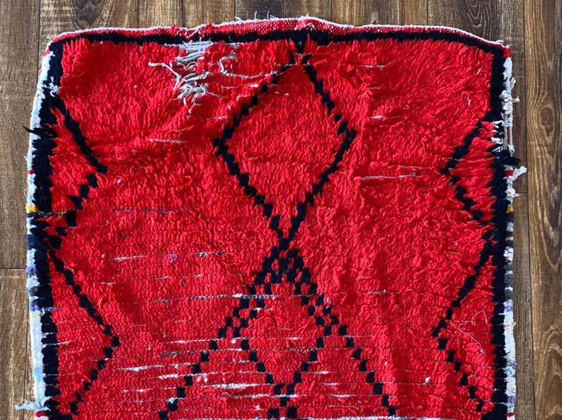 Op de afbeelding: Een rood Marokkaans tapijt met een zwart ruitpatroon. Het tapijt heeft een verweerde, vintage uitstraling en is gemaakt van wol.