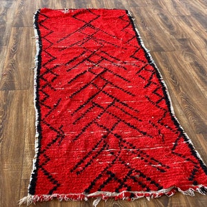 Op de afbeelding: Een rood en zwart Marokkaans tapijt met een ruitpatroon. Het tapijt heeft een gefranst randje en is gemaakt van wol.