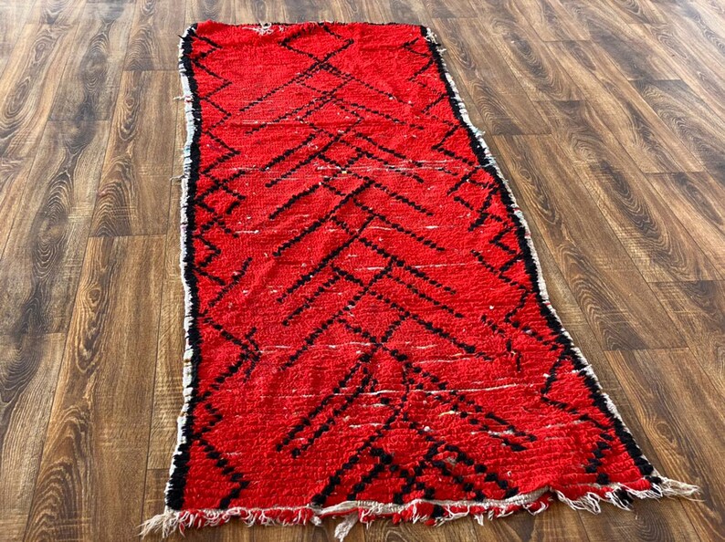 Op de afbeelding: Een rood en zwart Marokkaans tapijt met een ruitpatroon. Het tapijt heeft een gefranst randje en is gemaakt van wol.