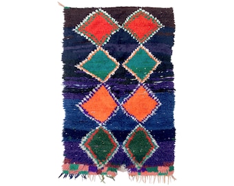 May include: A colorful Moroccan rug with a diamond pattern in shades of blue, green, orange, and red. The rug has a fringed edge.