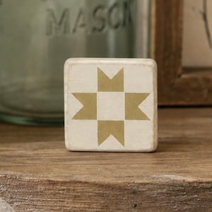 May include: A small square wooden block featuring a cream-colored surface with a gold-toned geometric star pattern. The block has a distressed finish on its edges and corners, presenting a simple, flat, and compact design for decorative display.