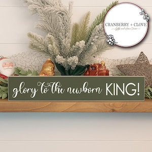 May include: A rectangular, olive green wooden sign with the text "glory to the newborn KING!" in white script. The sign is decorated with Christmas-themed elements, including greenery, ornaments, and a star.
