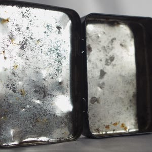 May include: Two empty metal tins with a worn, silver finish. The tins are rectangular and have a hinged lid.