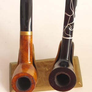 Puede incluir: Dos pipas de fumar de madera en un soporte de latón. Una pipa tiene un acabado negro con remolinos blancos y la otra tiene un acabado de madera natural con una banda de latón.