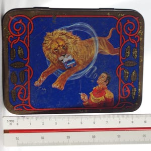 May include: A vintage metal tin with a blue background featuring a lion jumping through a smoke ring. A man in a red and gold uniform is looking up at the lion. The tin has a gold border with a red and gold design.